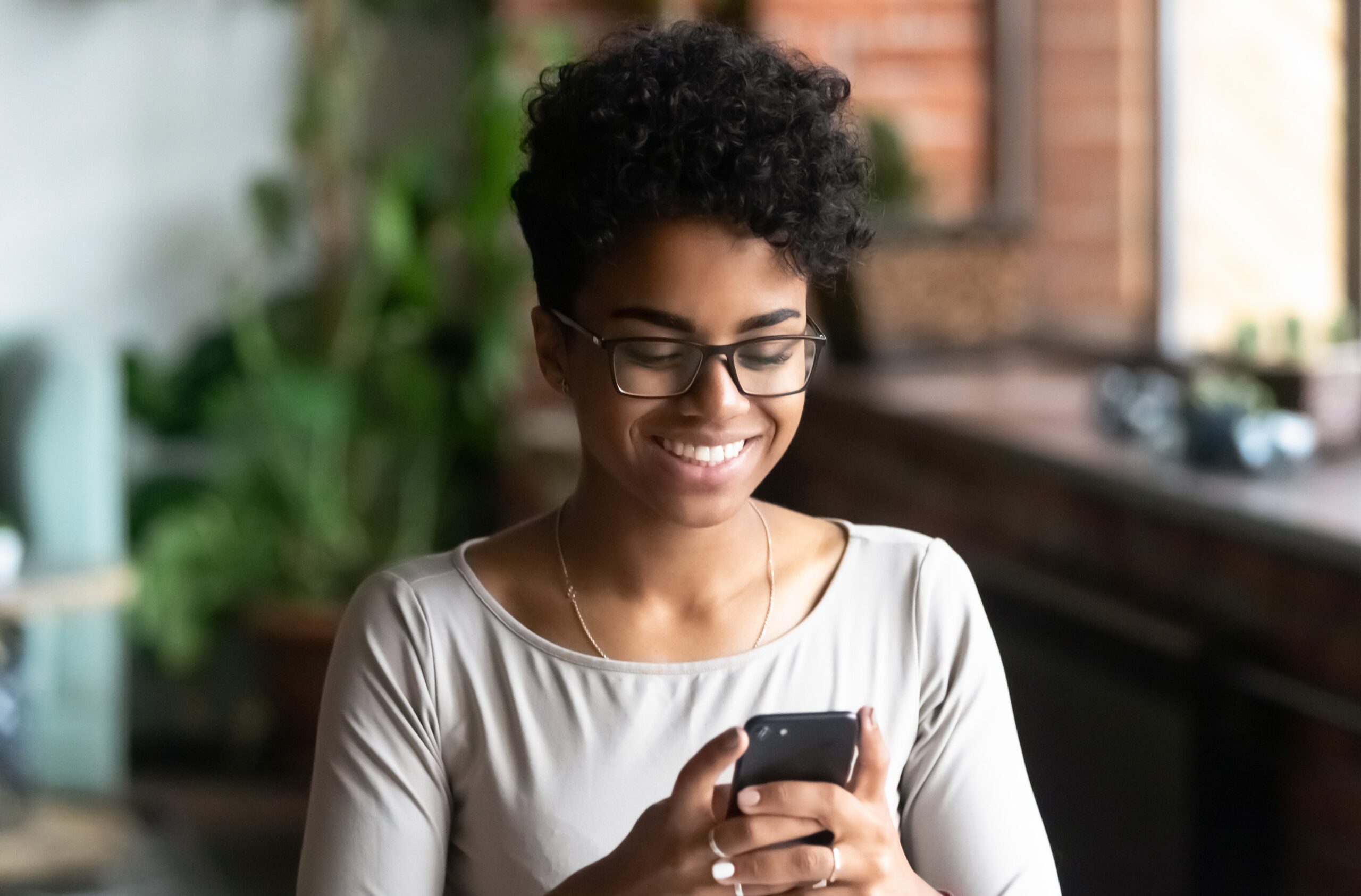 Cheerful african student black woman sitting at table studying using laptop reading a book, take a break holding mobile phone surfing internet received message from friend chatting about weekend plans